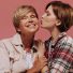 Mãe: símbolo de amor, força e determinação - Young short haired girl in plaid red shirt kissing on cheek her grandmother with blonde hair in beige jacket on pink backdrop.