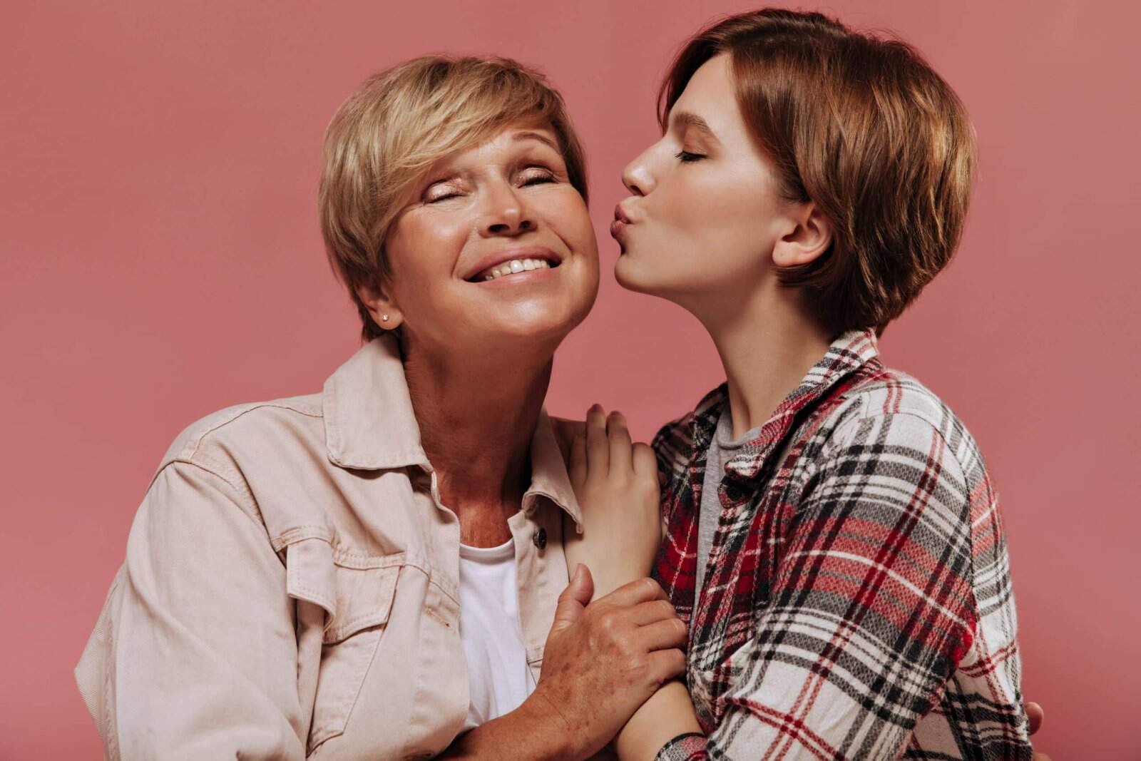 Mãe: símbolo de amor, força e determinação - Young short haired girl in plaid red shirt kissing on cheek her grandmother with blonde hair in beige jacket on pink backdrop.