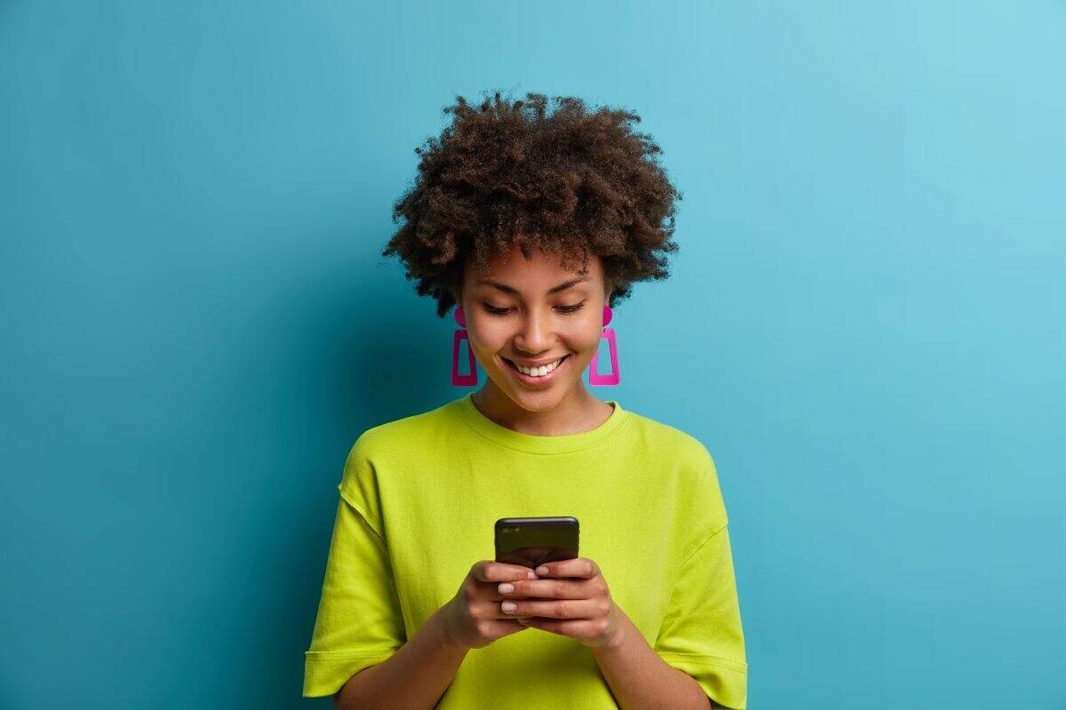Cheerful pleased woman with curly hair holds mobile phone and texts with friends in social networks, uses special application, watches interesting video, isolated on blue wall. People and technology