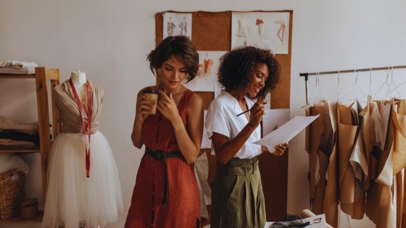Começando do Zero: guia para quem quer ser revendedora e nunca trabalhou com vendas 2 Duas mulheres em estúdio de moda, uma segurando um copo e a outra analisando um esboço, simbolizando o planejamento e os primeiros passos no modelo de revenda
