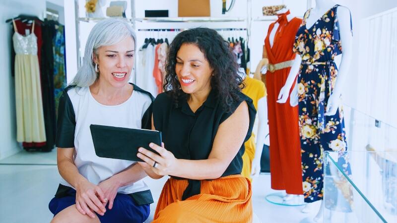 Começando do Zero: guia para quem quer ser revendedora e nunca trabalhou com vendas 3 Duas mulheres em loja de moda, olhando juntas para um tablet e sorrindo, representando a colaboração e o aprendizado nas primeiras vendas no modelo de revenda