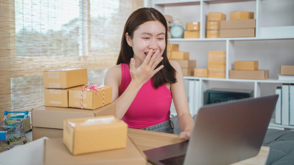 Mulher organizando pedidos e embalagens enquanto trabalha no notebook.